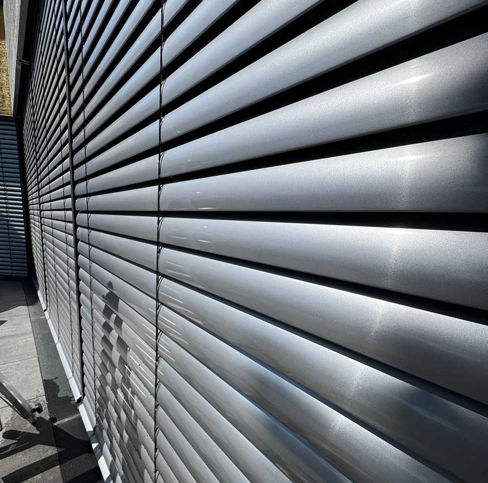 Metallische Jalousien in silberner Farbe, die in gleichmäßigen Abständen angeordnet sind und Licht reflektieren.