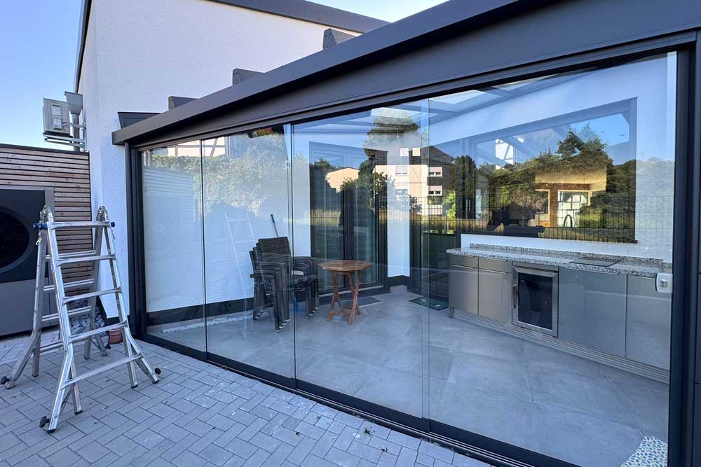 Moderne Glasveranda mit großen Fenstern, einem Tisch und Stühlen sowie einer Küchenzeile aus Edelstahl.