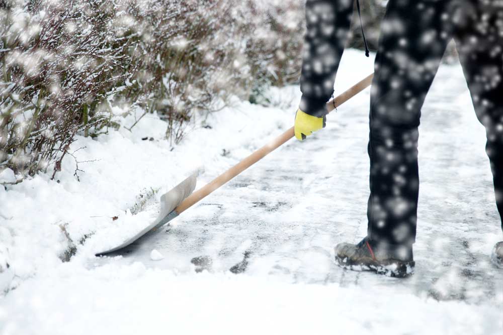 Person mit Schaufel, die Schnee von einem Gehweg entfernt, während es schneit. Handschuhe und robuste Kleidung sind sichtbar.